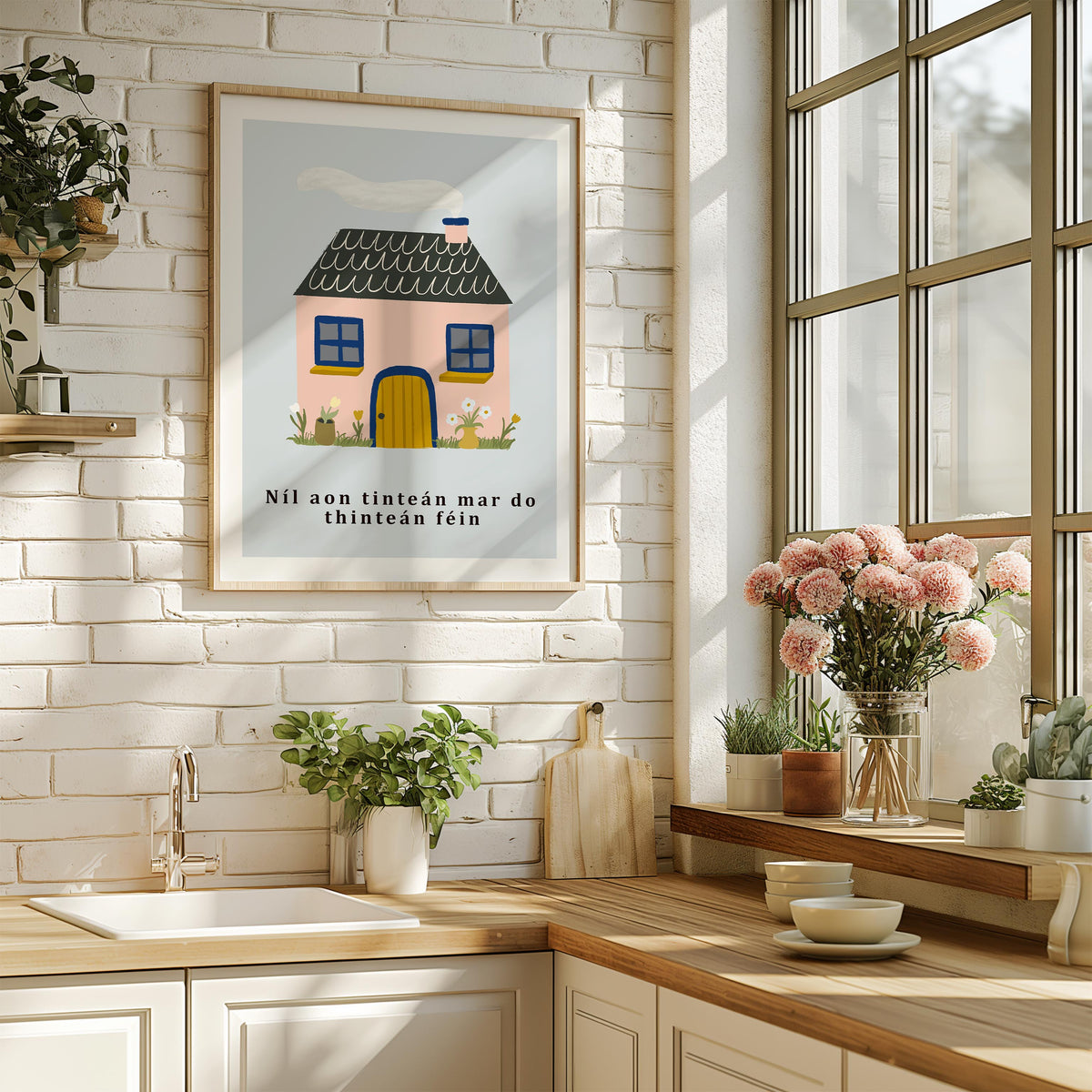 A framed art print featuring a whimsical pink cottage with a dark gray roof and the text 'Níl aon tinteán mar do thintéan fein' hangs on a white brick wall in a bright, modern kitchen with wooden countertops and potted plants.